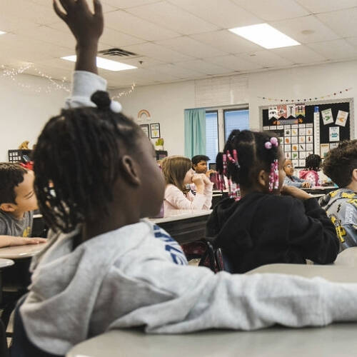 Students sitting at desks in a classroom, with a student in the foreground raising their hand.